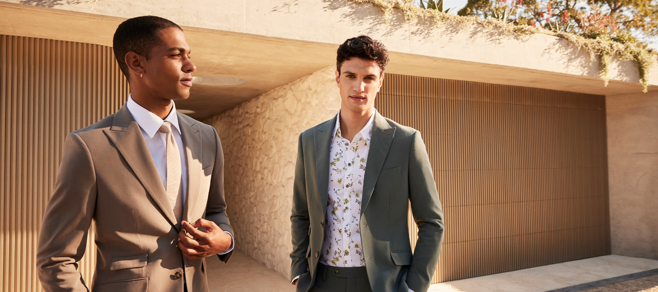 Two men wearing POLITIX Spring Summer Collection tailored suits – sage green suit with floral shirt and mushroom tailored suit – styled outdoors in modern architecture setting.